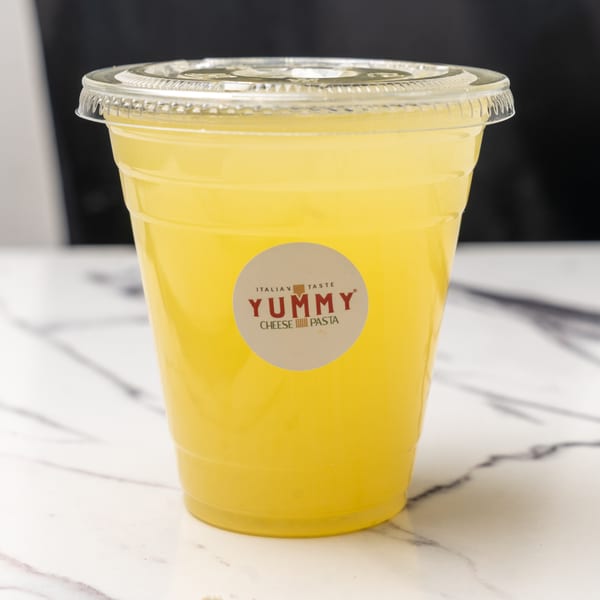 A clear plastic cup of yellow lemonade with a Yummy Cheese Pasta logo on a marble surface.
