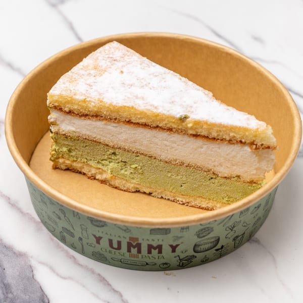 A slice of layered cake with green and white filling, topped with powdered sugar, in a Yummy branded container.